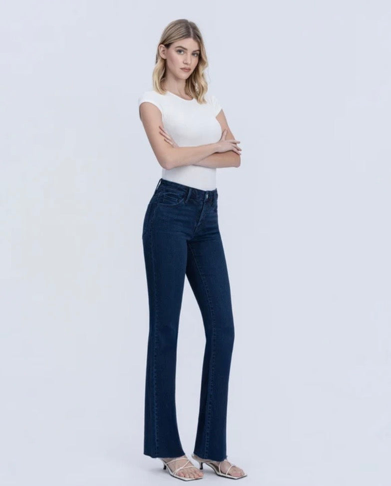 A woman modeling dark blue high-waisted flare jeans with a white sleeveless top, standing against a light gray studio background.