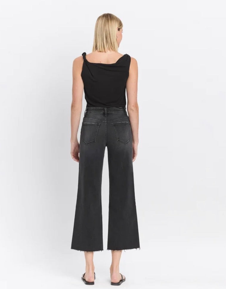 A woman shown from the back wearing a black draped sleeveless top and dark charcoal wide-leg cropped jeans with a frayed hem, standing against a plain white studio background.