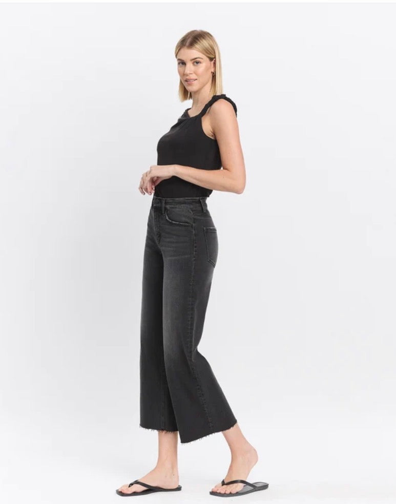 A woman modeling black wide-leg cropped jeans with a frayed hem, paired with a black sleeveless top and black flip-flops, against a plain white studio background.