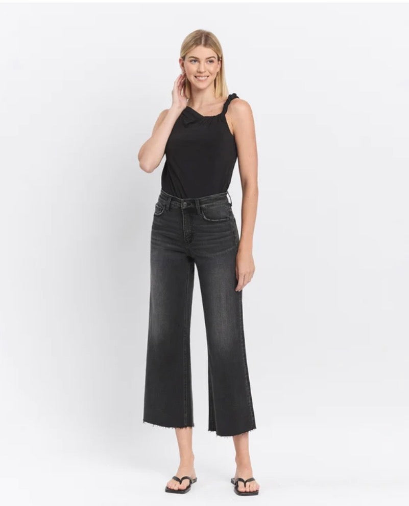 A woman modeling a black sleeveless top and dark charcoal wide-leg cropped jeans with frayed hems, paired with black flip-flop sandals, against a plain white studio background.