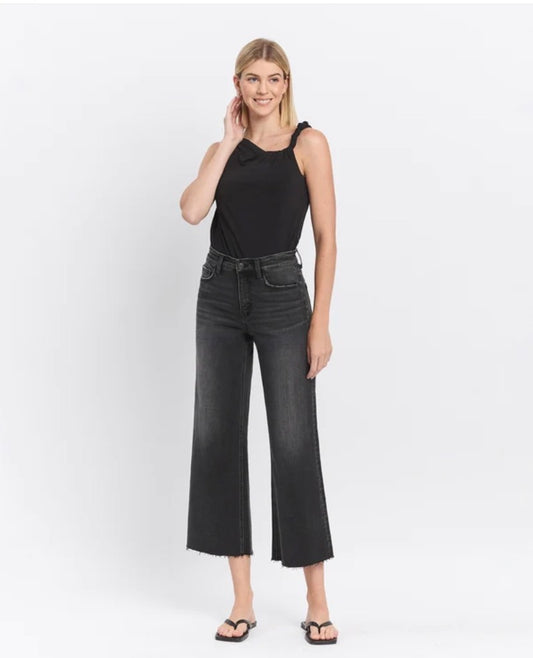A woman modeling a black sleeveless top and dark charcoal wide-leg cropped jeans with frayed hems, paired with black flip-flop sandals, against a plain white studio background.