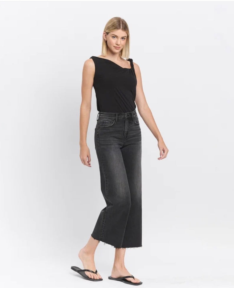 A woman modeling a black sleeveless top and dark gray cropped jeans against a plain white studio background.