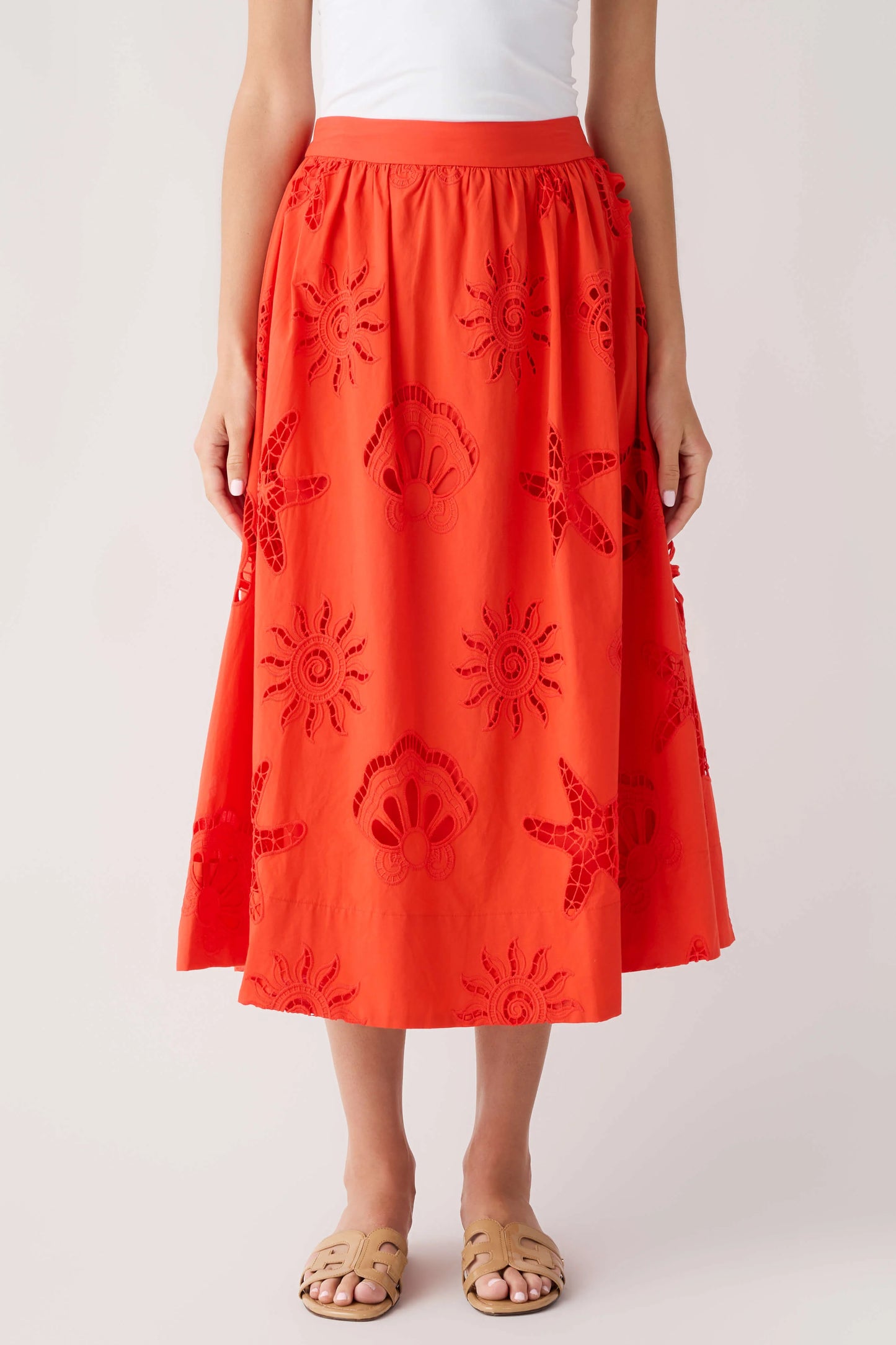 A model wearing a bright orange-red midi skirt with tonal embroidered sun, shell, and starfish motifs, paired with a white top and tan slide sandals, against a light gray studio background.