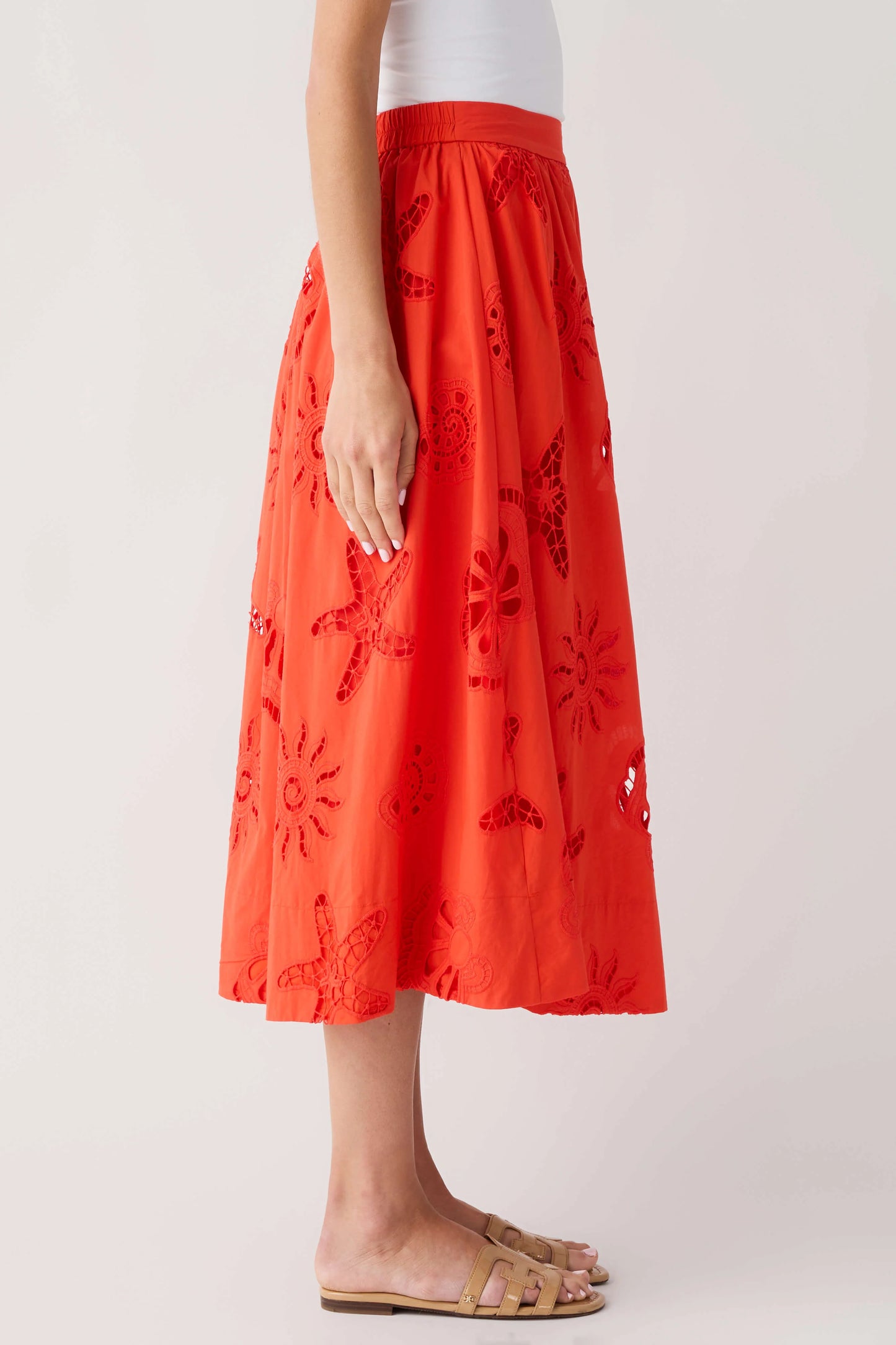 A model wearing a bright orange-red midi skirt with embroidered floral and butterfly patterns, styled with a white top and tan slide sandals against a light gray studio background.