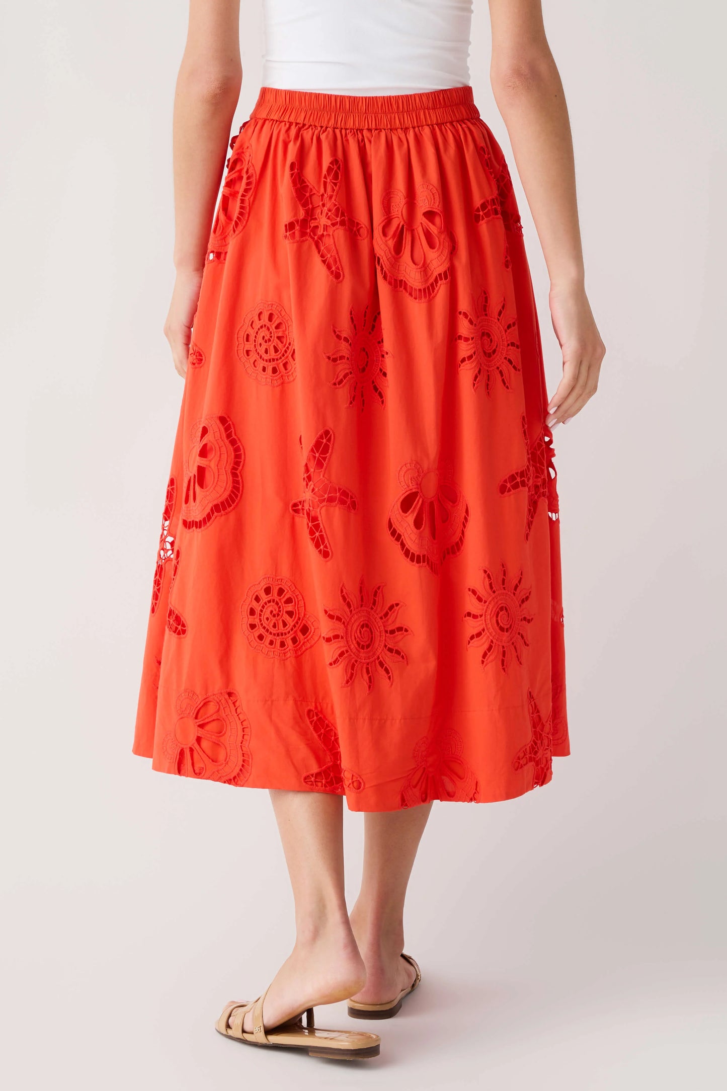 A model is shown from the waist down wearing a bright orange-red midi skirt with dark red embroidered floral and sunburst patterns, paired with a white top and tan slide sandals, against a light gray studio background.