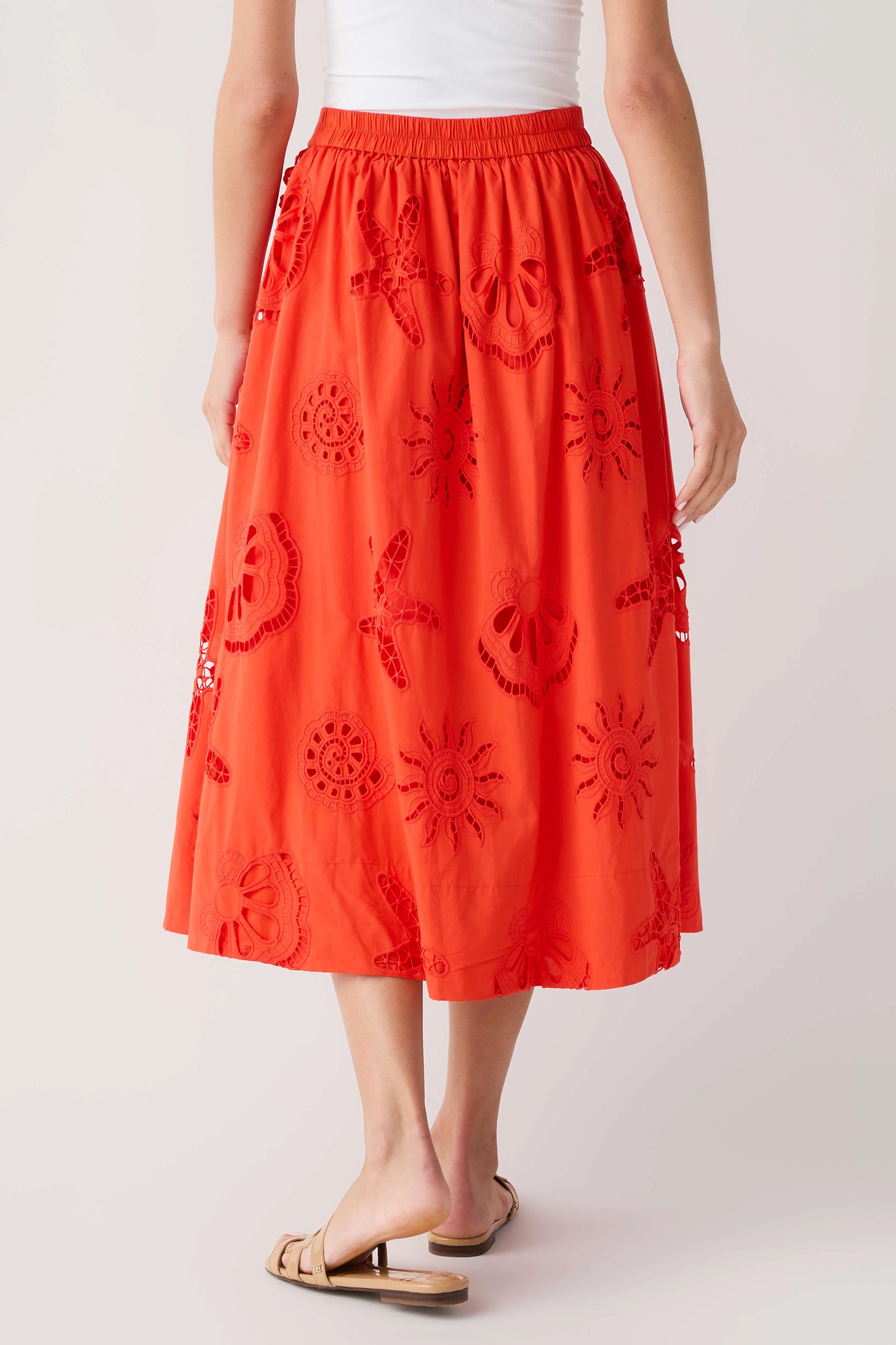 A model is shown from the waist down wearing a bright orange-red midi skirt with dark red embroidered floral and sunburst patterns, paired with a white top and tan slide sandals, against a light gray studio background.