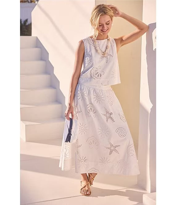 A woman models a sleeveless white maxi dress with tonal and raised starfish and shell embroidery, standing on white outdoor steps.