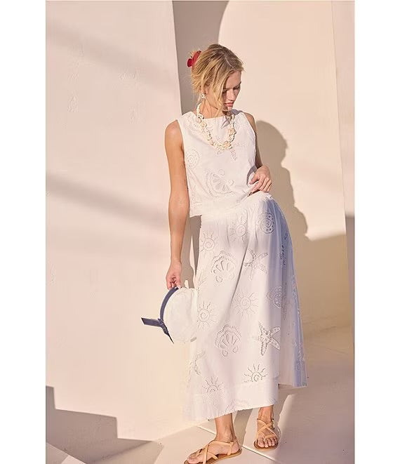 A woman modeling a sleeveless white maxi dress with tonal floral embroidery and a sheer light blue overlay skirt, standing in a minimalist beige room with strong sunlight and shadows.