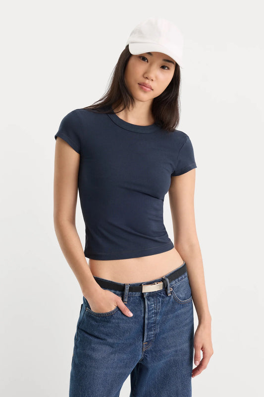 A woman modeling a navy blue short-sleeve crop top with a white baseball cap and blue jeans against a plain light gray background.