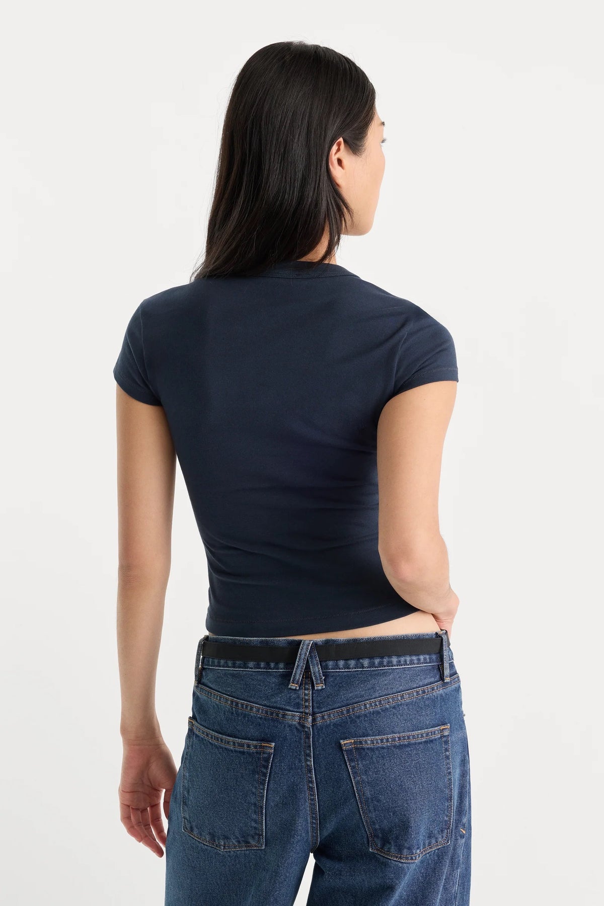 A woman viewed from the back wearing a dark navy blue short-sleeve crop top and blue jeans against a plain white studio background.