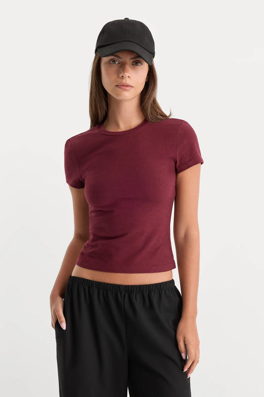A woman modeling a fitted short-sleeve crewneck crop top in deep burgundy, paired with black sweatpants and a black baseball cap, against a plain light gray studio background.
