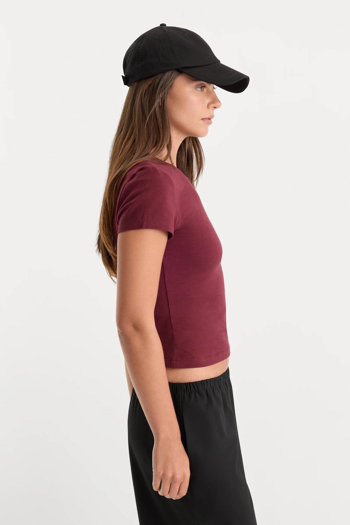 A woman in side profile wearing a short-sleeve maroon crop top, black pants, and a black baseball cap against a plain light gray background.