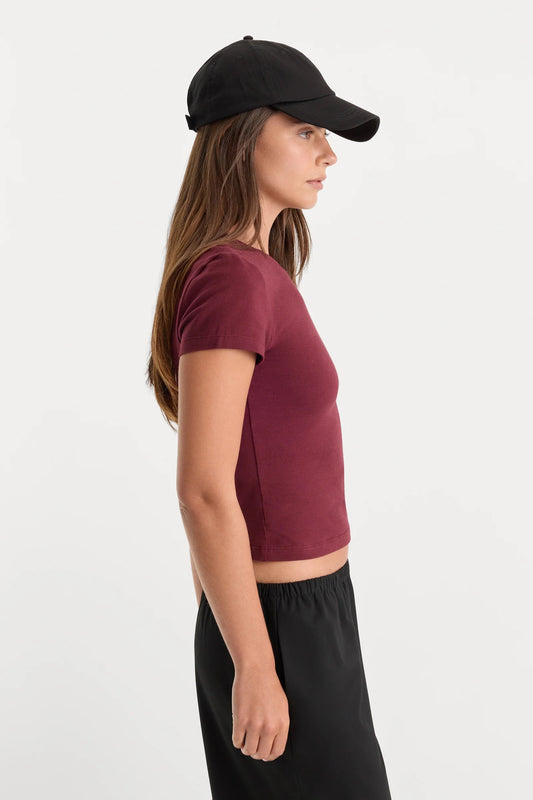 A woman in side profile wearing a short-sleeve maroon crop top, black pants, and a black baseball cap against a plain light gray background.