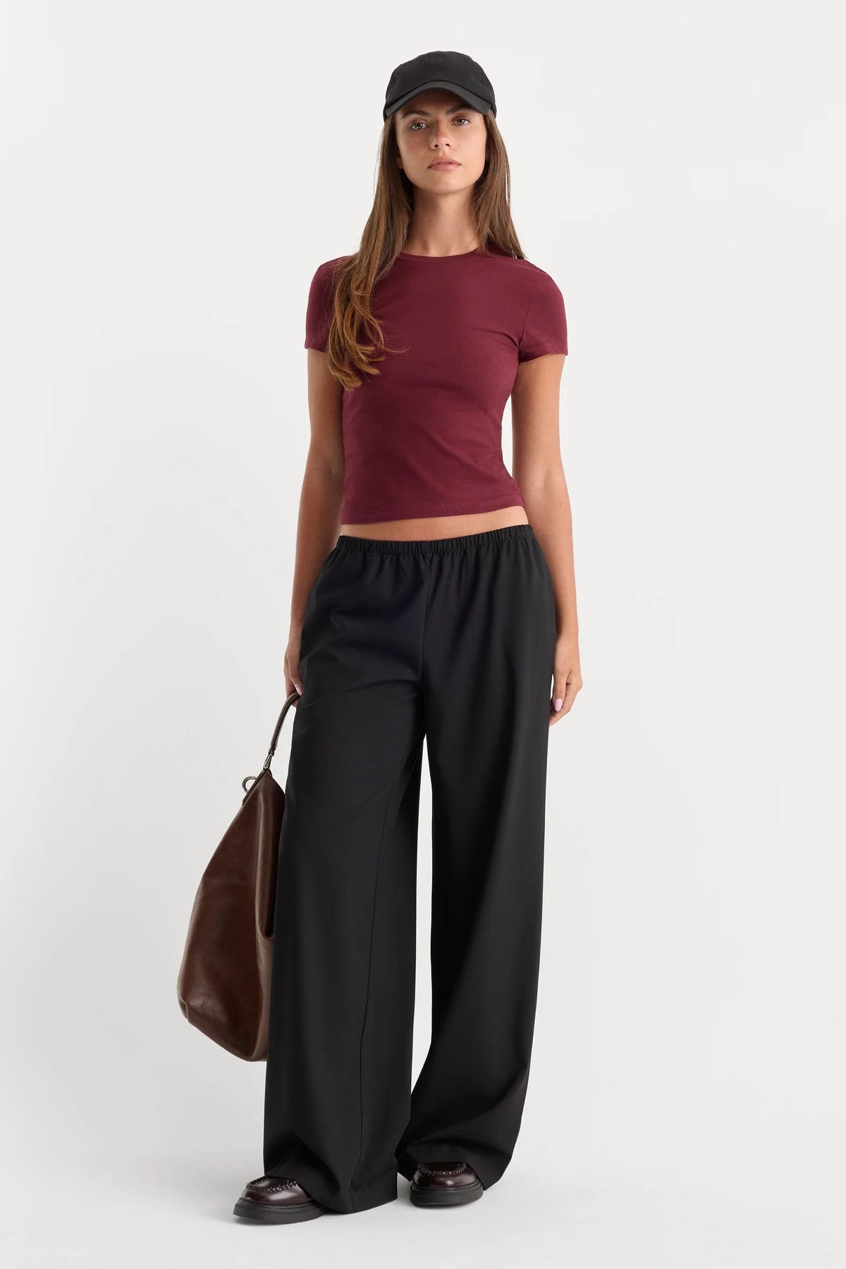 A woman modeling a fitted short-sleeve burgundy crop top, black wide-leg pants, a black baseball cap, and carrying a brown leather tote bag against a light gray studio background.