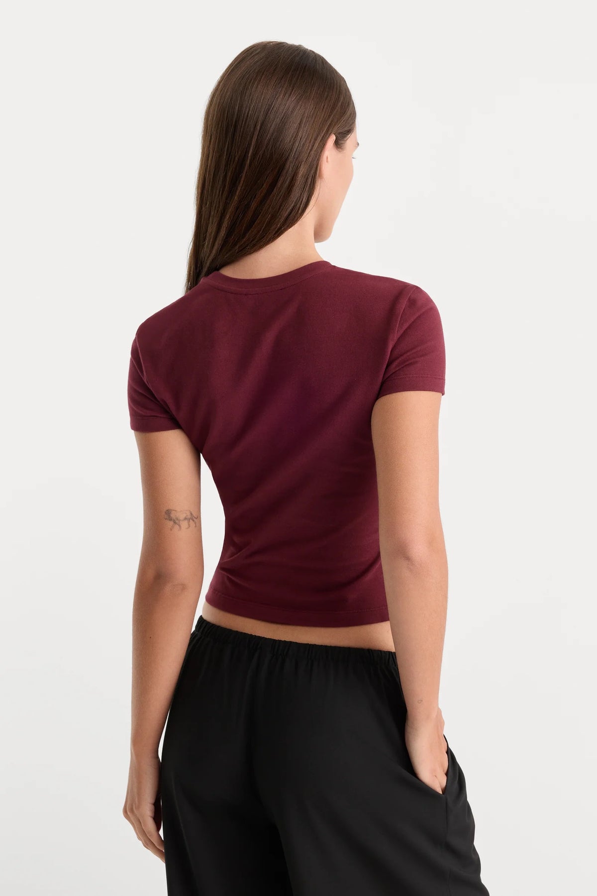 A woman shown from the back wearing a short-sleeve maroon crop top and black pants against a plain white studio background.
