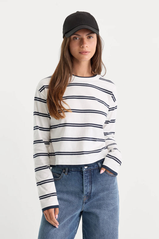 A woman wearing a white long-sleeve crewneck sweater with thin navy horizontal stripes, paired with blue jeans and a black baseball cap, photographed against a plain light gray studio background.