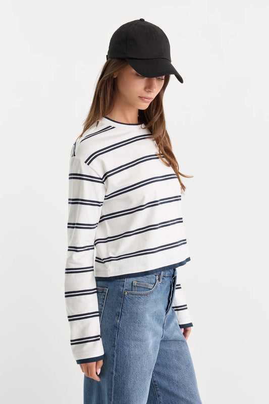 A model wearing a white long-sleeve cropped t-shirt with navy blue horizontal stripes, paired with blue jeans and a black baseball cap, photographed against a plain white studio background.