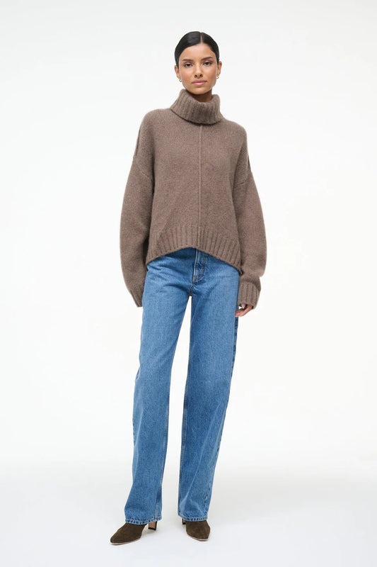 Person wearing a brown turtleneck sweater and blue jeans on a white background