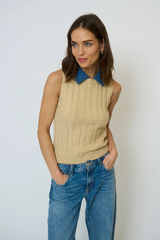 Woman wearing a beige sleeveless sweater with a blue collar and blue jeans against a white background