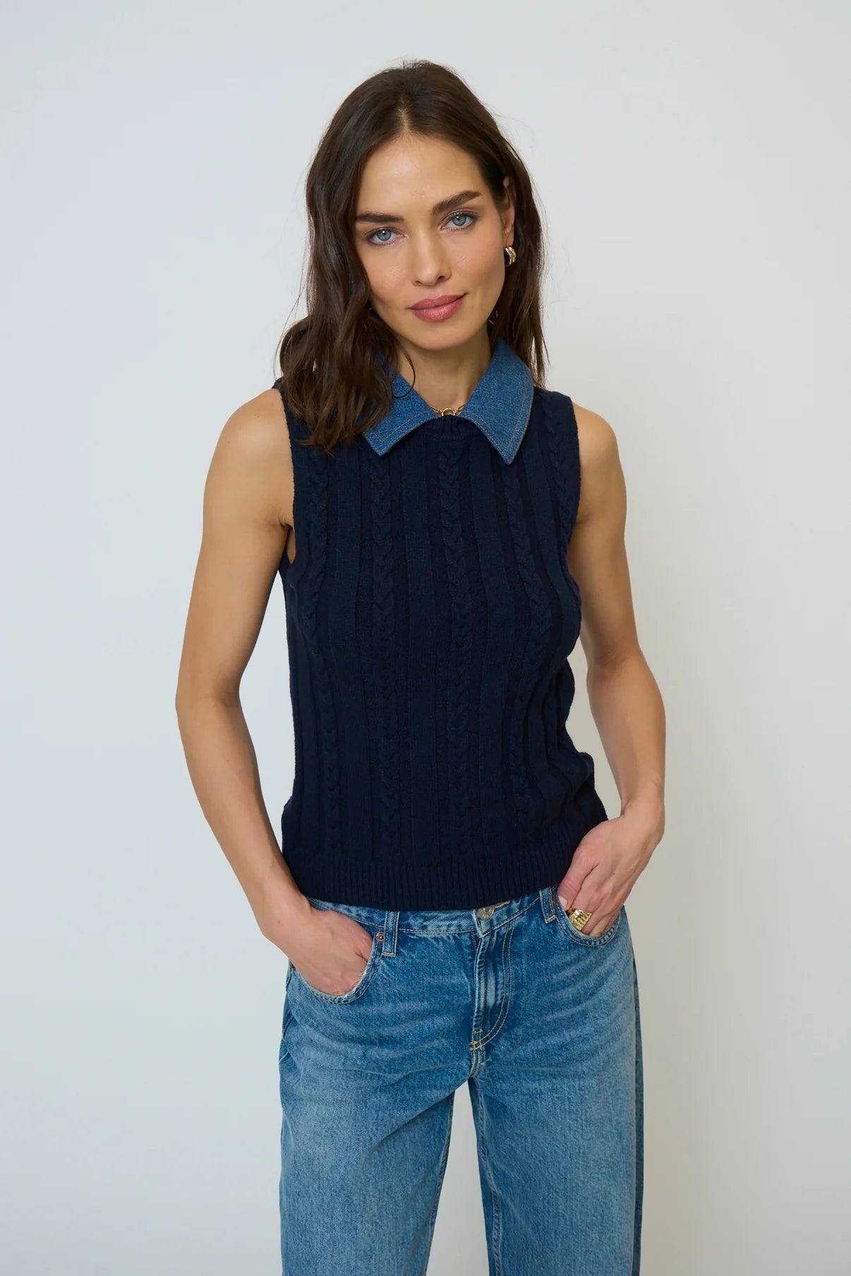 Woman wearing a navy sleeveless sweater with a blue collar and jeans against a white background