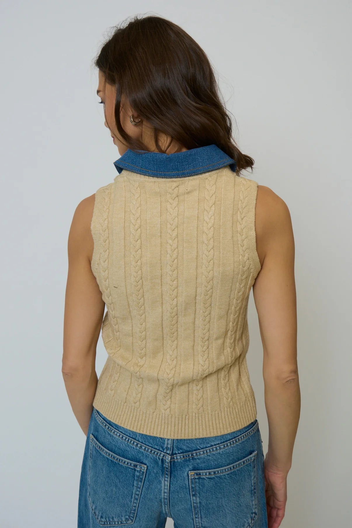 Woman wearing a beige sleeveless knitted sweater and blue jeans against a plain background