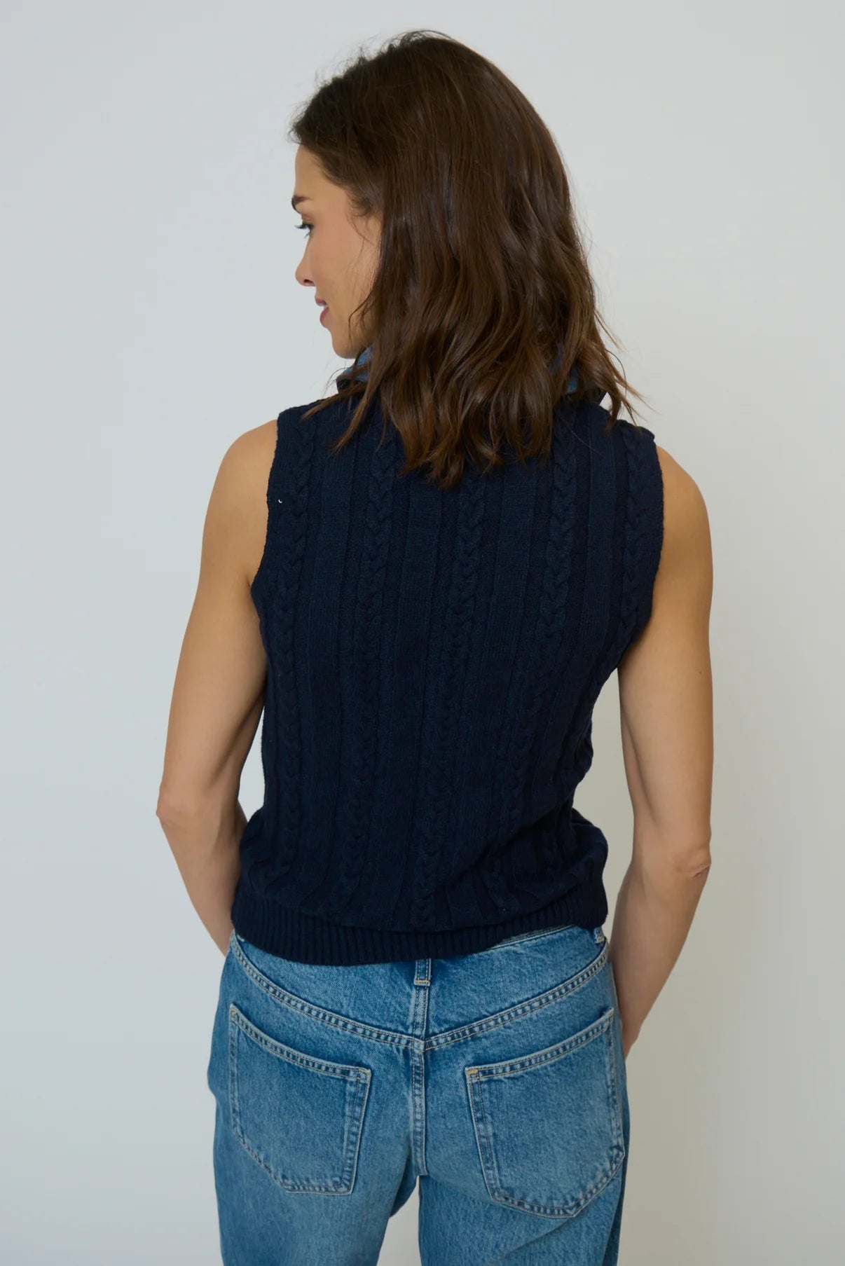 Woman wearing a navy sleeveless sweater and blue jeans against a white background
