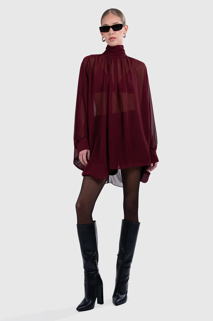Person wearing a burgundy dress with sheer panels and black knee-high boots on a white background