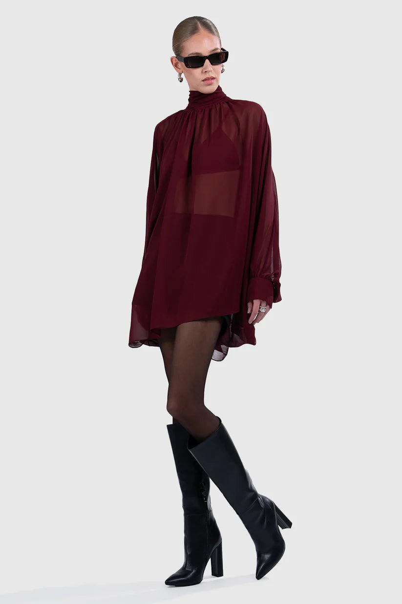Woman wearing a burgundy dress with sheer panels and black knee-high boots on a white background