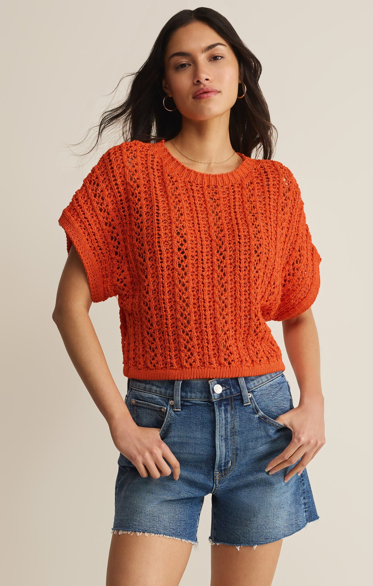 Woman wearing an orange knitted top and denim shorts on a beige background