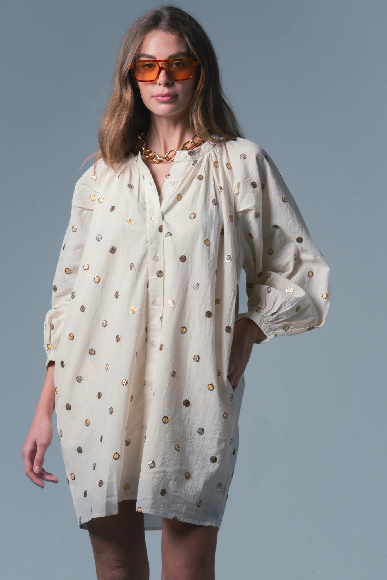 Woman wearing a white dress with gold polka dots and sunglasses on a gray background