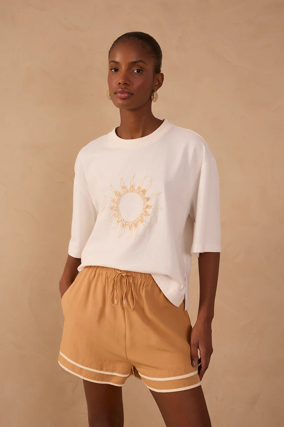 Woman wearing a white sweater with a sun design and beige shorts on a beige background