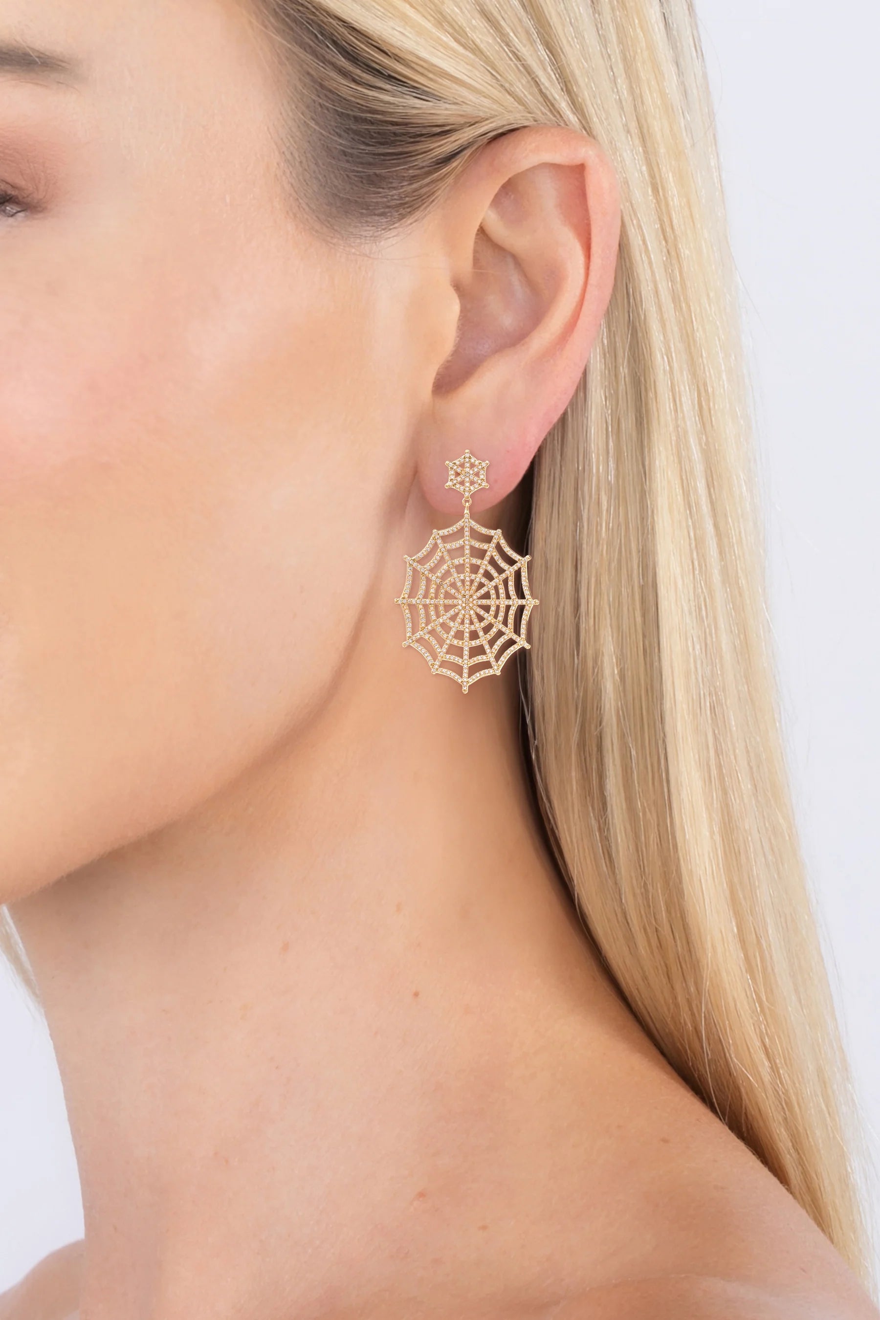 A close-up of a woman's ear wearing a delicate gold spiderweb dangle earring against a clean white background.