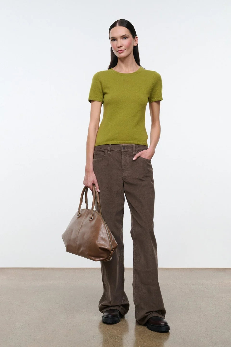 A woman modeling a short-sleeve crewneck knit top in olive green, paired with brown pants, against a plain light gray studio background.