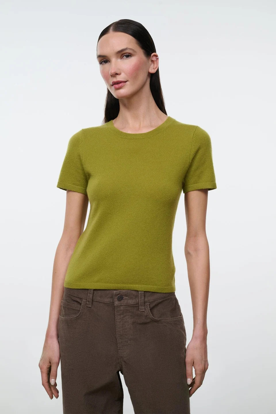 A woman modeling a short-sleeve crewneck knit top in olive green, paired with brown pants, against a plain light gray studio background.