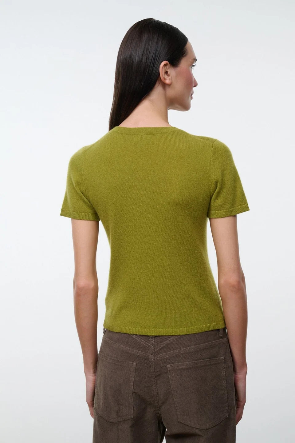 A woman shown from the back wearing a short-sleeve olive green knit top and brown pants, photographed against a plain light gray studio background.