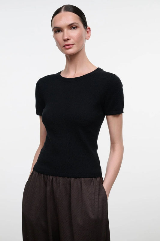 A woman modeling a short-sleeve crewneck knit top in solid black, paired with dark brown wide-leg trousers, against a clean light gray studio background.