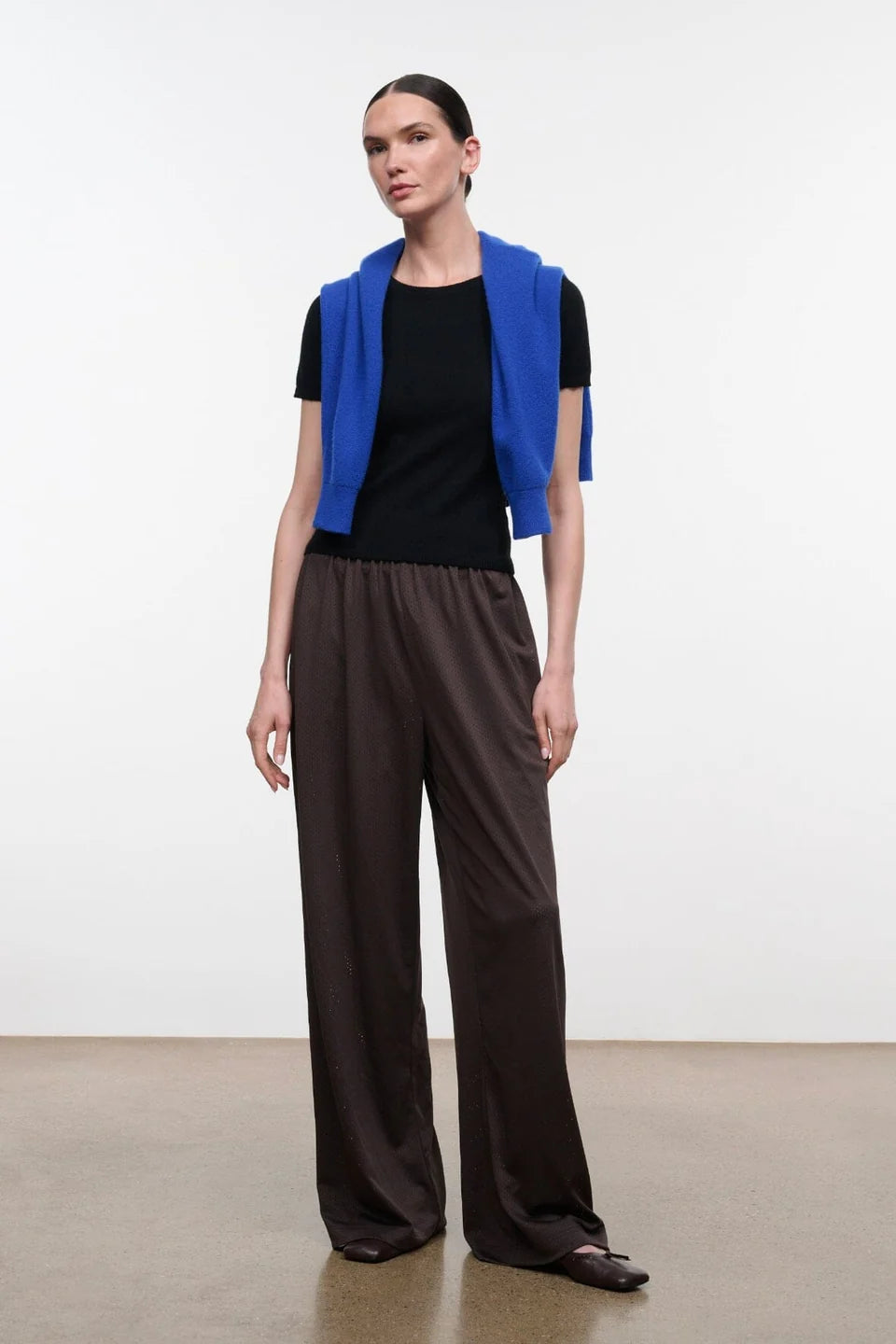 A model stands in a studio wearing a bright blue sleeveless vest over a black short-sleeve top and dark brown wide-leg trousers.