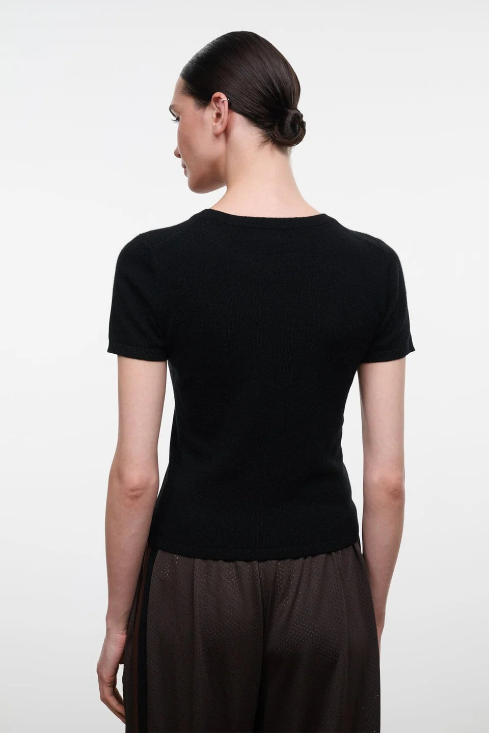 A woman shown from the back wearing a fitted black short-sleeve top and dark brown pleated trousers, photographed against a plain light gray studio background.
