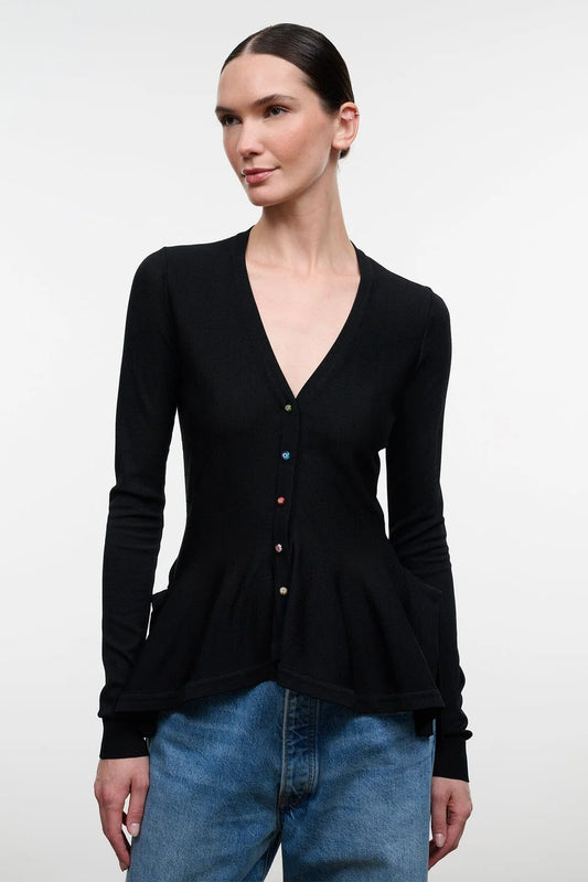 A woman modeling a black long-sleeve button-front top with a peplum hem, paired with wide-leg blue jeans and dark sandals, standing against a plain light gray studio background.
