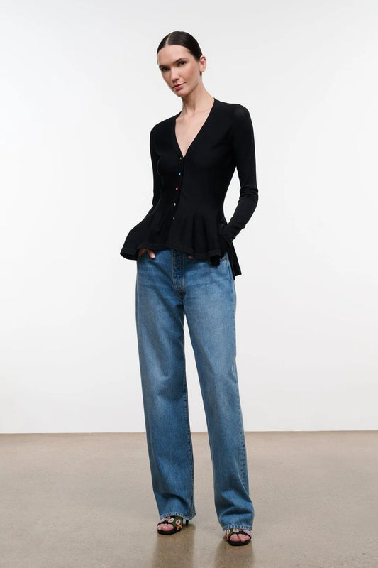 A woman modeling a black long-sleeve button-front top with a peplum hem, paired with wide-leg blue jeans and dark sandals, standing against a plain light gray studio background.