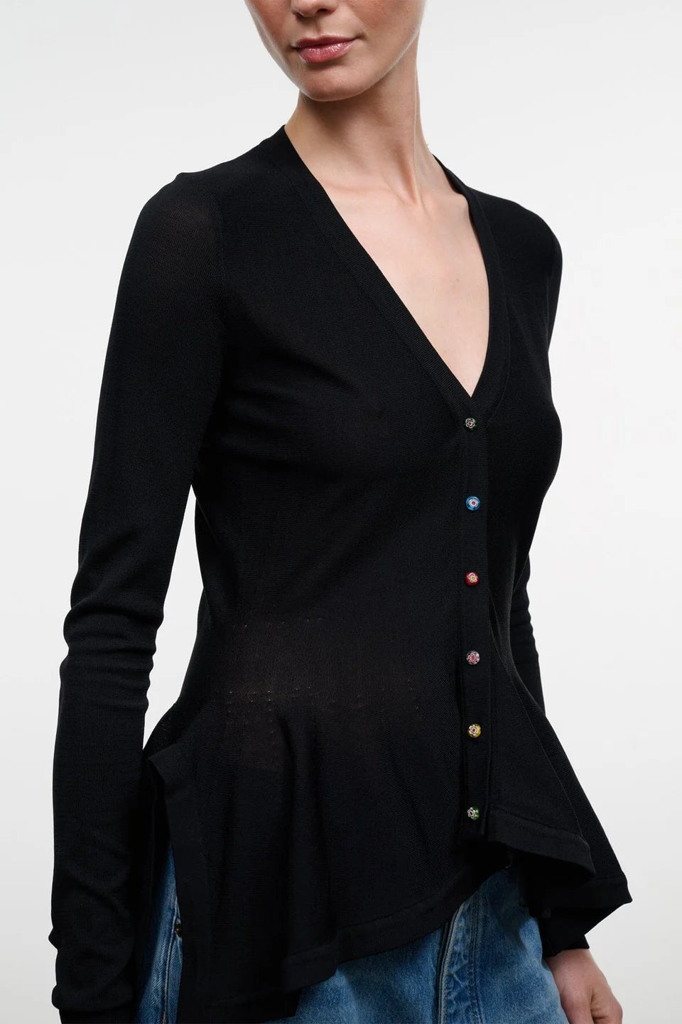A woman wearing a black long-sleeve V-neck button-front top with colorful buttons, paired with blue jeans, against a plain light gray background.