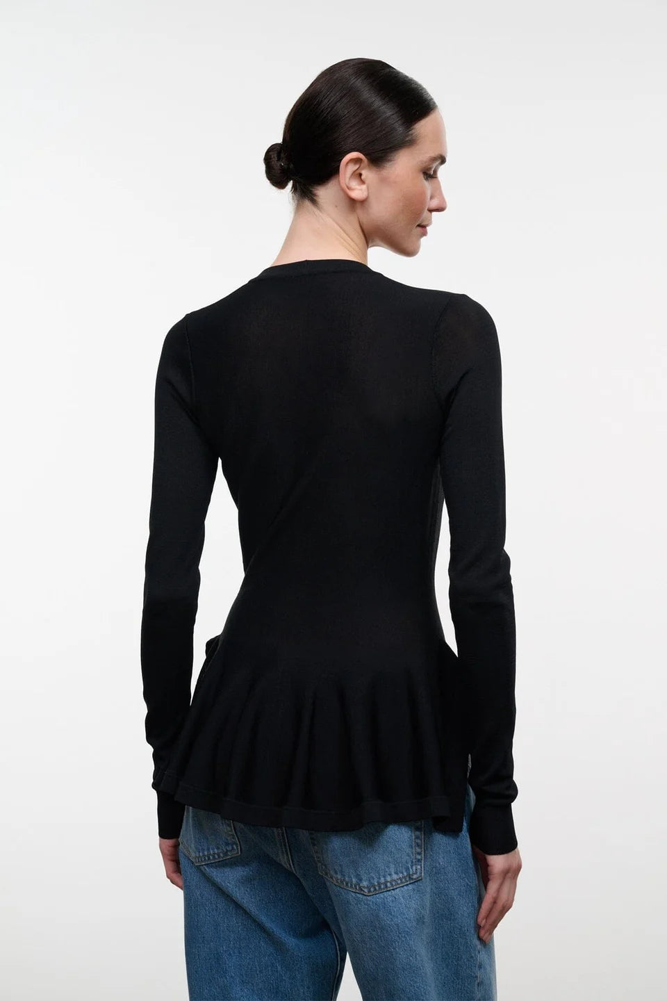 The back of a woman modeling a black long-sleeve top with a peplum hem, paired with wide-leg blue jeans and dark sandals, standing against a plain light gray studio background.