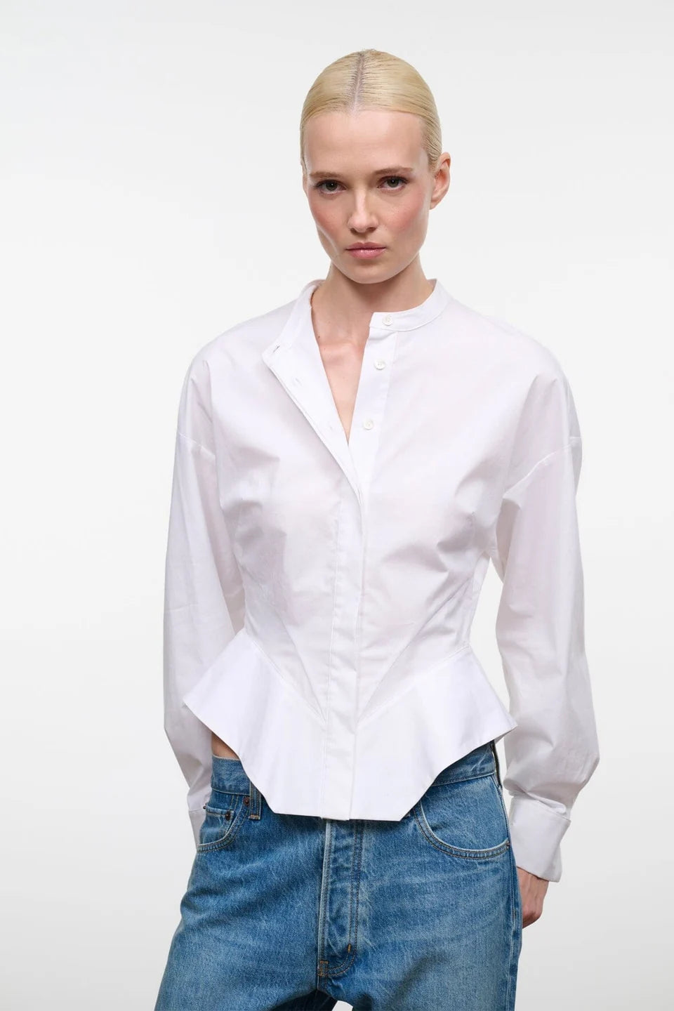 A blonde model wearing a white long-sleeve button-up shirt with a split collar and a peplum hem, paired with blue jeans, against a plain white studio background.