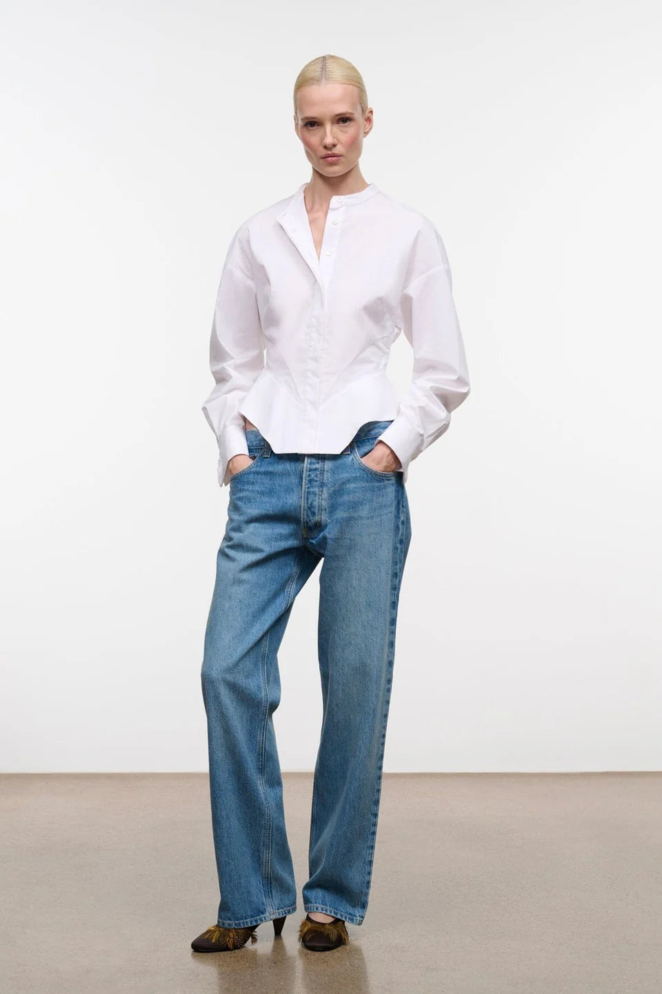 A model stands in a studio wearing a white long-sleeve button-up shirt tucked into blue wide-leg jeans, paired with brown pointed shoes, against a plain white wall and beige floor.