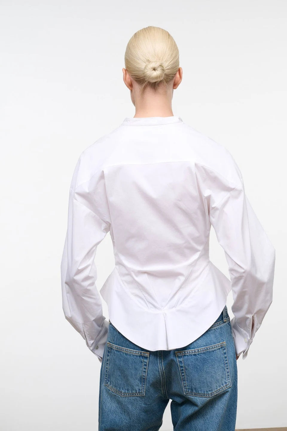 A person viewed from behind wearing a white long-sleeve button-up shirt and blue jeans against a plain light gray studio background.
