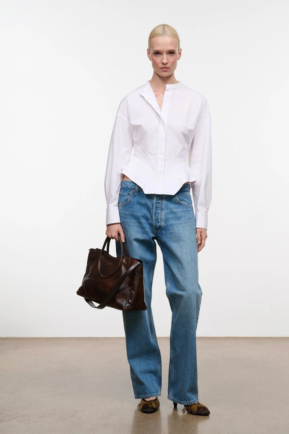 A model stands in a studio wearing a white long-sleeve button-up shirt, blue wide-leg jeans, and brown shoes, while holding a dark brown leather tote bag.