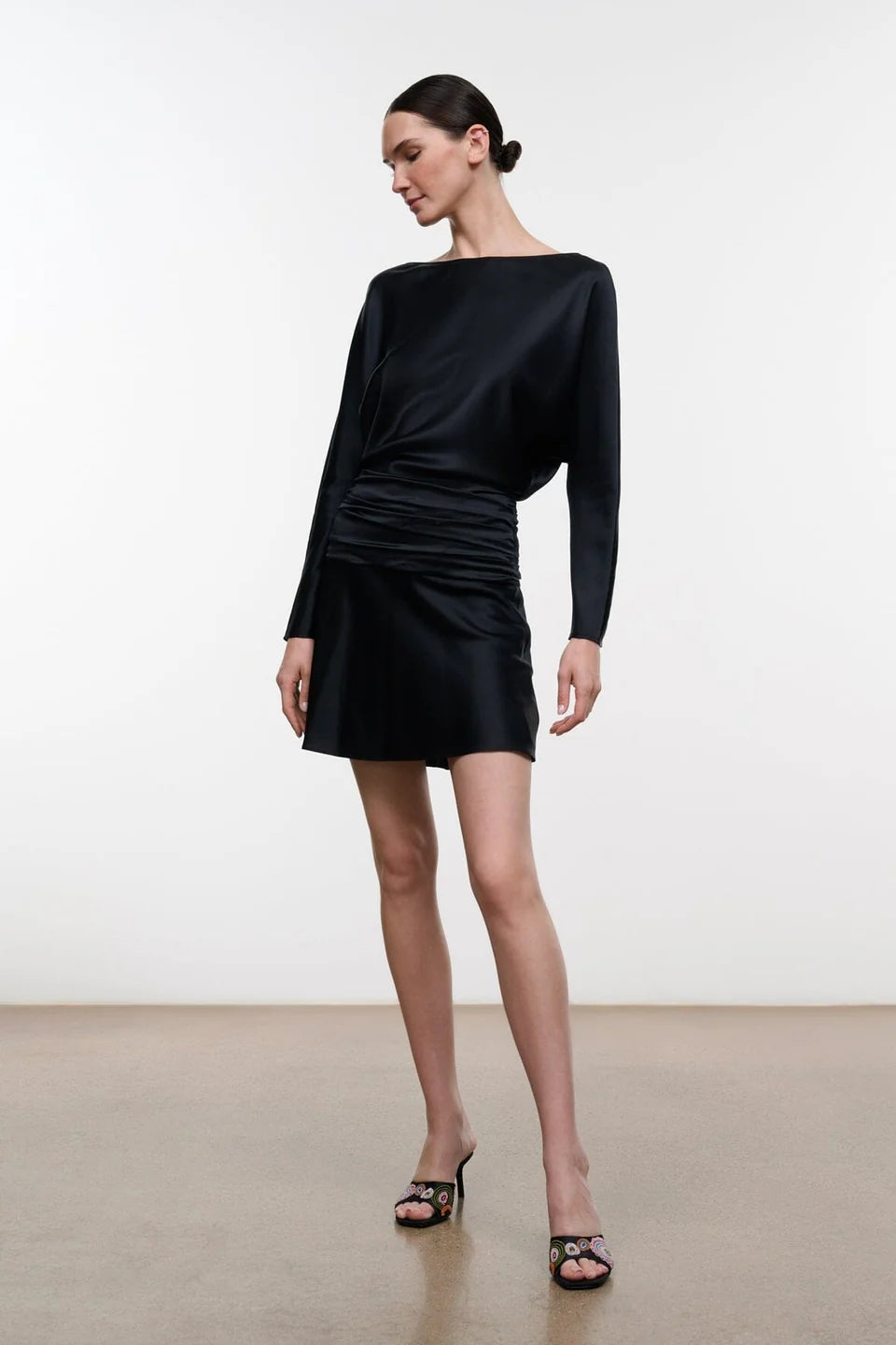 A model wearing a black long-sleeve mini dress with a draped, gathered waist and boat neckline, paired with black strappy heeled sandals, set against a plain white studio background.