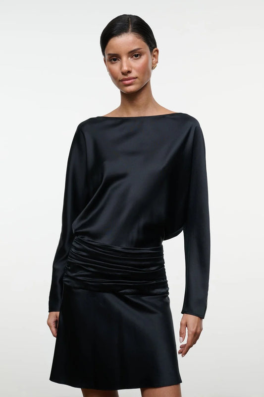 A woman modeling a black satin long-sleeve mini dress with a draped, asymmetrical neckline and gathered ruching at the waist, set against a plain light gray studio background.