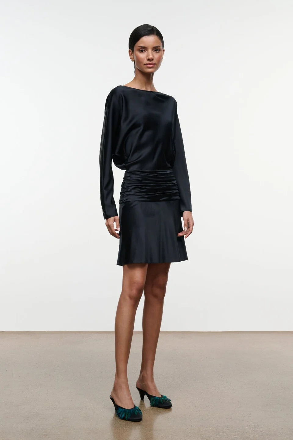 A model wearing a black satin long-sleeve mini dress with a gathered ruched bodice and flared skirt, paired with dark green velvet mule heels, set against a minimalist studio background.