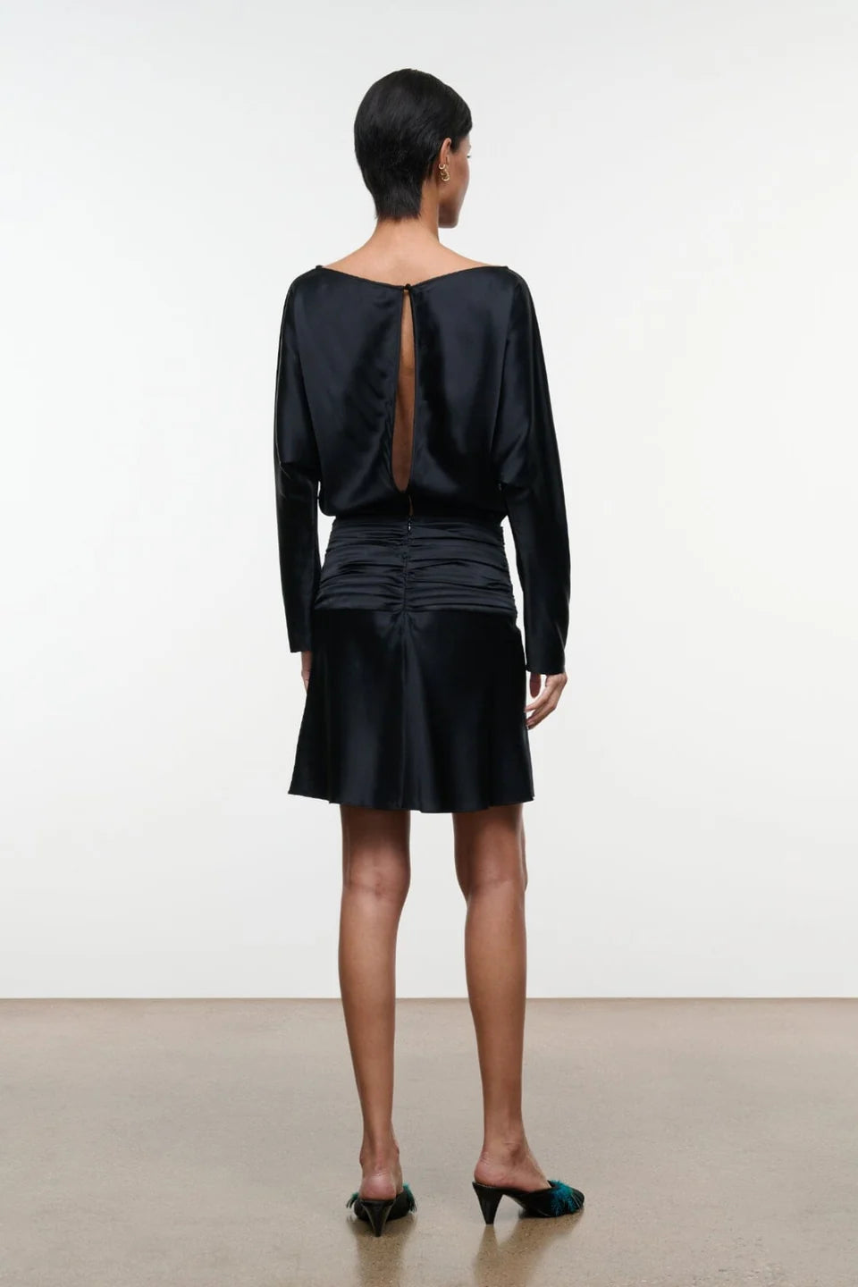 A model viewed from the back wearing a black satin long-sleeve dress with a dramatic open back and gathered skirt, paired with dark green mule heels, set against a minimalist studio background.